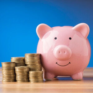 a smiling, from on, pink piggy bank next to some piles of gold coins on a wooden bench with a blue background