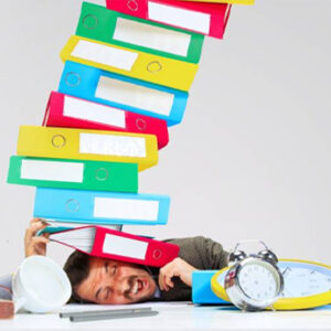 a man with his head laying on a desk. on top of his head is pile many different colored binder folders. The man looks weighed down by the folders and his facial expression is a grimace. There is a tipped over mug and an analog alarm clock on the desk in front of the man