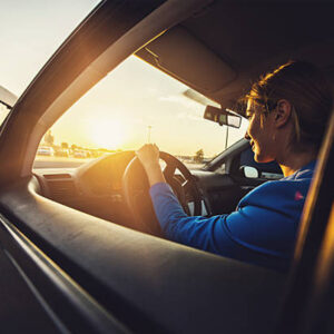 a closeup photo of a person driving a car taken through the drivers side open window. The sun is shining through the windscreen