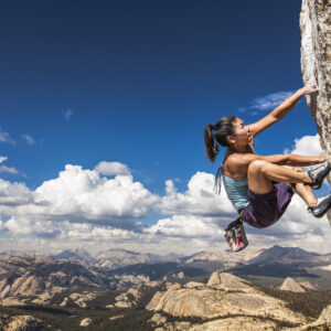 a side view of a woman rock climbing a rock wall. In the distance is a blue sky with clouds and mountains